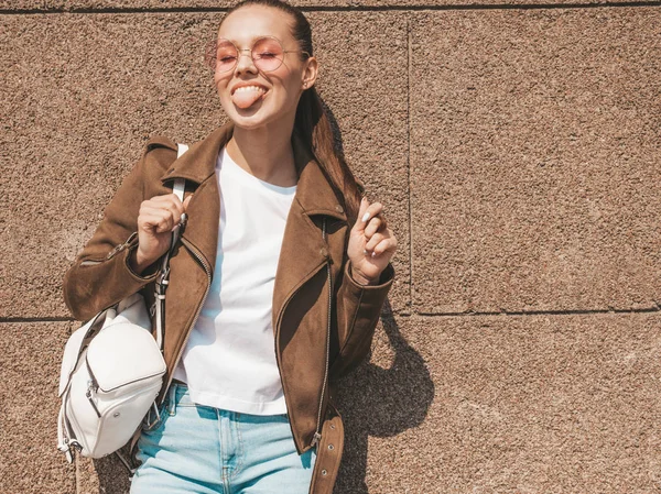 Yaz hipster ceket giysiler giymiş güzel gülümseyen esmer model portresi. Duvara yakın sokakta poz veren modaya uygun kız. Yuvarlak güneş gözlüğü komik ve pozitif kadın. Dilini göstermek