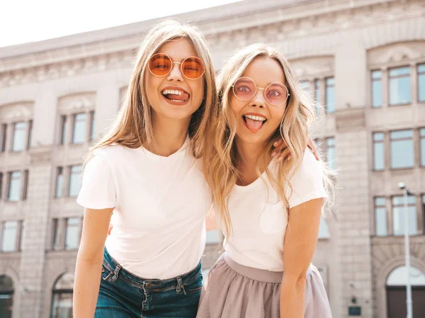 两个年轻美丽的金发微笑的嬉皮士女孩在时髦的夏季白色T恤衣服的肖像 性感的无忧无虑的女人在街头背景摆姿势 正面模型显示他们的舌头 在太阳镜 — 图库照片