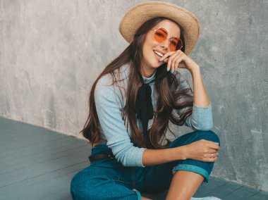 Genç, güzel, gülümseyen bir kadın kameraya bakıyor. Günlük kıyafetleri ve şapkası olan modaya uygun bir kız. Stüdyoda gri duvarın yanında poz veren komik ve pozitif bir kadın. Yerde oturan güneş gözlükleriyle