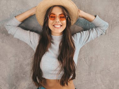 Genç, güzel, gülümseyen bir kadın kameraya bakıyor. Sıradan yaz kıyafetleri ve şapkası olan modaya uygun bir kız. Stüdyoda gri duvarın yanında poz veren komik ve pozitif kadın.