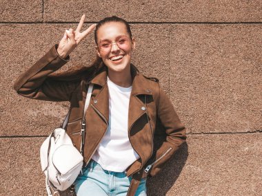 Beautiful smiling brunette model dressed in summer hipster jacket and jeans clothes. Trendy girl posing in the street near wall. Funny and positive woman in round sunglasses showing peace sign