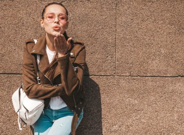Beautiful smiling brunette model dressed in summer hipster jacket and jeans clothes. Trendy girl posing in the street near wall. Funny and positive woman in round sunglasses giving air kiss