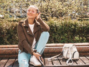 Yaz hipster ceket ve kot elbise giymiş güzel gülümseyen esmer model Portresi. Sokak arka planda bankta oturan trendy kız. Yuvarlak güneş gözlüğü komik ve pozitif kadın