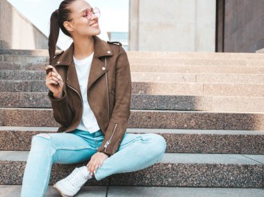 Yaz hipster ceket ve kot elbise giymiş güzel gülümseyen esmer model Portresi. Sokak arka planda merdivenlerde oturan trendy kız. Yuvarlak güneş gözlüğü komik ve pozitif kadın