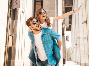 Gülümseyen güzel kız ve rahat yazlık giysiler onun yakışıklı erkek arkadaşı. Kız arkadaşını sırtında taşıyan adam ellerini kaldırıyor. Mutlu neşeli aile sokak arka planda eğlenmek 