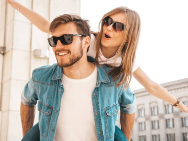 Gülümseyen güzel kız ve rahat yazlık giysiler onun yakışıklı erkek arkadaşı. Kız arkadaşını sırtında taşıyan adam ellerini kaldırıyor. Mutlu neşeli aile sokak arka planda eğlenmek 