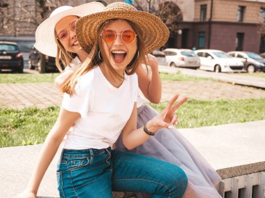 Trendy yaz beyaz t-shirt giysiler iki genç güzel sarışın gülümseyen hipster kız. Seksi kaygısız kadınlar sokak arka planda poz. Pozitif modeller güneş gözlüğü ve şapka eğlenmek. Barış işareti gösterme