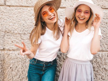 Trendy yaz beyaz t-shirt giysiler iki genç güzel sarışın gülümseyen hipster kız. Duvarın yanındaki sokakta poz veren kadınlar. Pozitif modeller güneş gözlüğü ve şapka eğlenmek. Barış işareti gösterme