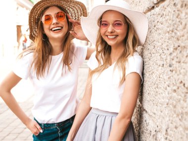 Moda yaz beyaz t-shirt giysiler iki genç güzel sarışın gülümseyen hipster kız Portresi. Seksi kaygısız kadınlar duvara yakın sokakta poz. Güneş gözlüğü ve şapka ile eğlenen pozitif modeller