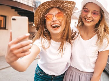 İki genç, gülümseyen, beyaz tişörtlü sarışın kadın. Akıllı telefondan selfie çeken kızlar. Arka planda poz veren mankenler. Olumlu yüz ifadeleri gösteren bir kadın.