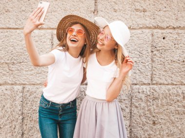 İki genç, gülümseyen, beyaz tişörtlü sarışın kadın. Akıllı telefondan selfie çeken kızlar. Arka planda poz veren mankenler. Olumlu yüz ifadeleri gösteren bir kadın.
