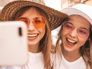 İki genç, gülümseyen, beyaz tişörtlü sarışın kadın. Akıllı telefondan selfie çeken kızlar. Arka planda poz veren mankenler. Olumlu yüz ifadeleri gösteren bir kadın.