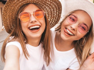 İki genç, gülümseyen, beyaz tişörtlü sarışın kadın. Akıllı telefondan selfie çeken kızlar. Arka planda poz veren mankenler. Olumlu yüz ifadeleri gösteren bir kadın.