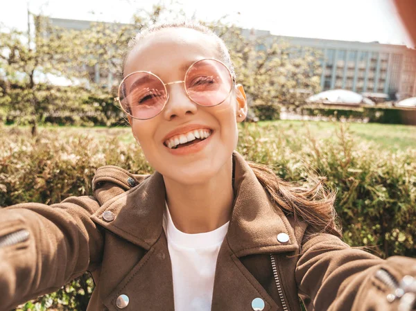 Beautiful smiling girl in summer hipster jacket and jeans.Model taking selfie on smartphone.Woman making photos in the street. Sitting on the bench in sunglasses