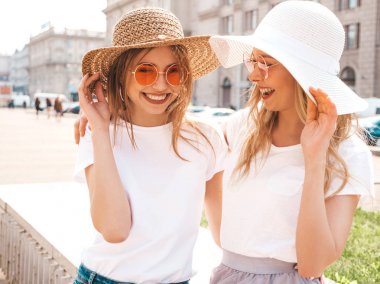 Moda yaz beyaz t-shirt giysiler iki genç güzel sarışın gülümseyen hipster kız Portresi. Seksi kaygısız kadınlar sokak arka planda poz. Güneş gözlüğü ve şapka ile eğlenen pozitif modeller