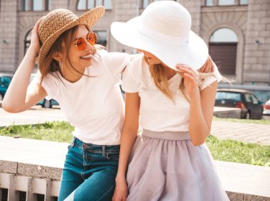 Moda yaz beyaz t-shirt giysiler iki genç güzel sarışın gülümseyen hipster kız Portresi. Seksi kaygısız kadınlar sokak arka planda poz. Güneş gözlüğü ve şapka ile eğlenen pozitif modeller