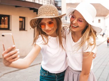 İki genç, gülümseyen, beyaz tişörtlü sarışın kadın. Akıllı telefondan selfie çeken kızlar. Arka planda poz veren mankenler. Olumlu yüz ifadeleri gösteren bir kadın.