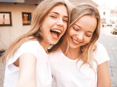 İki genç, gülümseyen, beyaz tişörtlü sarışın kadın. Akıllı telefondan selfie çeken kızlar. Arka planda poz veren mankenler. Olumlu yüz ifadeleri gösteren bir kadın.