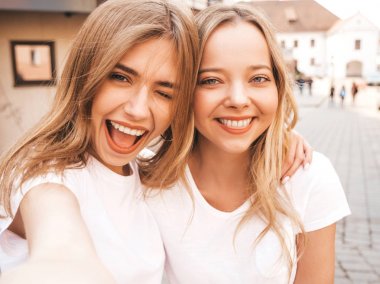İki genç, gülümseyen, beyaz tişörtlü sarışın kadın. Akıllı telefondan selfie çeken kızlar. Arka planda poz veren mankenler. Olumlu yüz ifadeleri gösteren bir kadın.