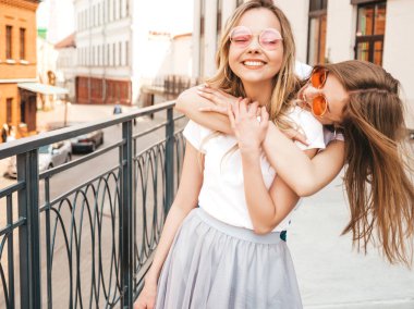 Moda yaz beyaz t-shirt giysiler iki genç güzel sarışın gülümseyen hipster kız Portresi. Seksi kaygısız kadınlar sokak arka planda poz. Güneş gözlüğü ile eğlenen pozitif modeller. Sarılma