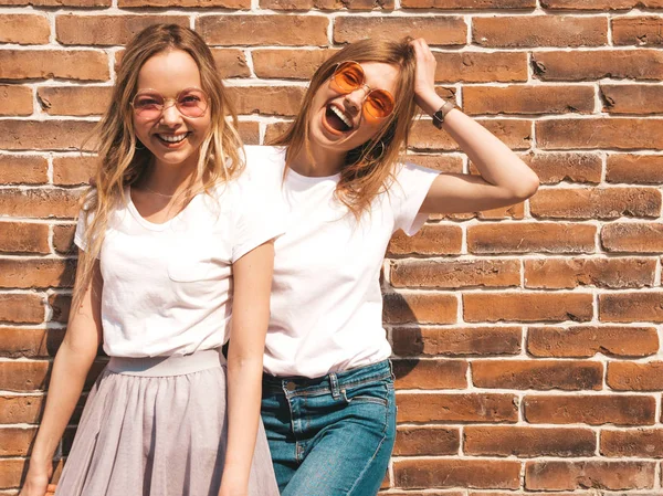 トレンディな夏の白いTシャツの服を着た2人の若い美しいブロンドの笑顔ヒップスターの女の子 レンガの壁の近くの通りでポーズをとる女性たち サングラスで楽しむポジティブなモデル — ストック写真