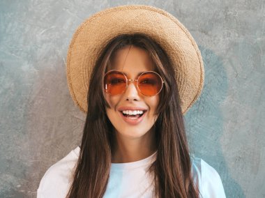 Genç, güzel, gülümseyen bir kadın kameraya bakıyor. Gündelik tişört ve şapka giyen modaya uygun bir kız. Stüdyoda güneş gözlüklü, gri duvarın yanında poz veren komik ve pozitif kadın.. 