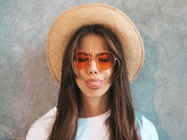 Genç, güzel, gülümseyen bir kadın kameraya bakıyor. Gündelik tişört ve şapka giyen modaya uygun bir kız. Stüdyoda güneş gözlükleriyle gri duvarın yanında poz veren komik ve pozitif bir kadın.