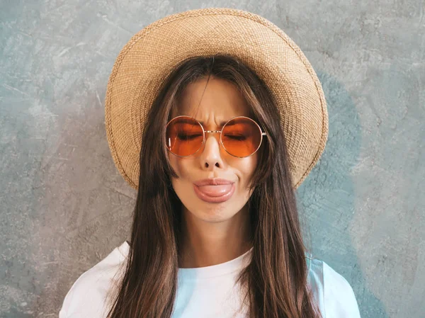 Genç, güzel, gülümseyen bir kadın kameraya bakıyor. Gündelik tişört ve şapka giyen modaya uygun bir kız. Stüdyoda güneş gözlükleriyle gri duvarın yanında poz veren komik ve pozitif bir kadın.