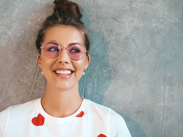 Kameraya bakan genç güzel gülümseyen kadının yakın çekim portresi. Rahat yaz beyaz elbise ve güneş gözlüğü trendy kız. Komik ve pozitif kadın stüdyoda gri duvar yakınında poz