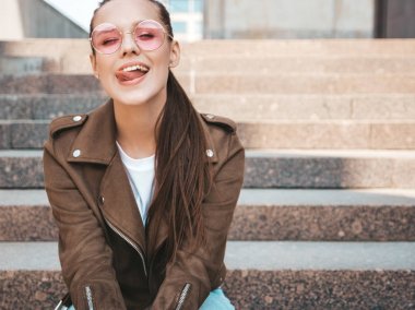 Yaz hipster ceket giysiler giymiş güzel gülümseyen esmer model portresi. Sokak arka planda poz veren modaya uygun bir kız. Yuvarlak güneş gözlüğü komik ve pozitif kadın. Dili gösterme