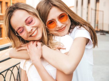 Moda yaz beyaz t-shirt giysiler iki genç güzel sarışın gülümseyen hipster kız Portresi. Seksi kaygısız kadınlar sokak arka planda poz. Güneş gözlüğü ile eğlenen pozitif modeller. Sarılma