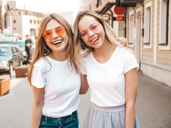 两个年轻美丽的金发微笑的嬉皮士女孩在时髦的夏季白色T恤衣服的肖像 性感的无忧无虑的女人在街头背景摆姿势 正款车型在太阳镜中玩得开心 — 图库照片