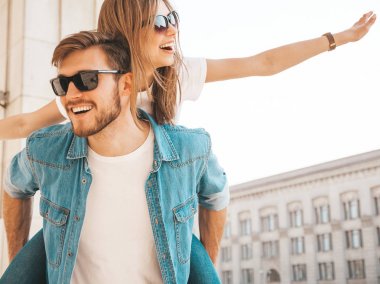 Gülümseyen güzel kız ve rahat yazlık giysiler onun yakışıklı erkek arkadaşı. Kız arkadaşını sırtında taşıyan adam ellerini kaldırıyor. Mutlu neşeli aile sokak arka planda eğlenmek 