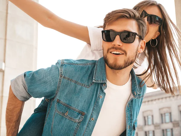 Smiling beautiful girl and her handsome boyfriend in casual summer clothes. Man carrying his girlfriend on the back and she raising her hands.Happy cheerful family having fun on the street background