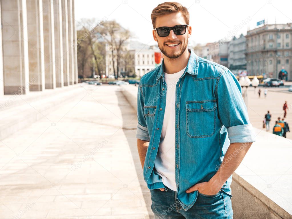 Portrait of handsome smiling stylish hipster lumbersexual businessman model. Man dressed in jeans jacket clothes. Fashion male posing on the street background in sunglasses