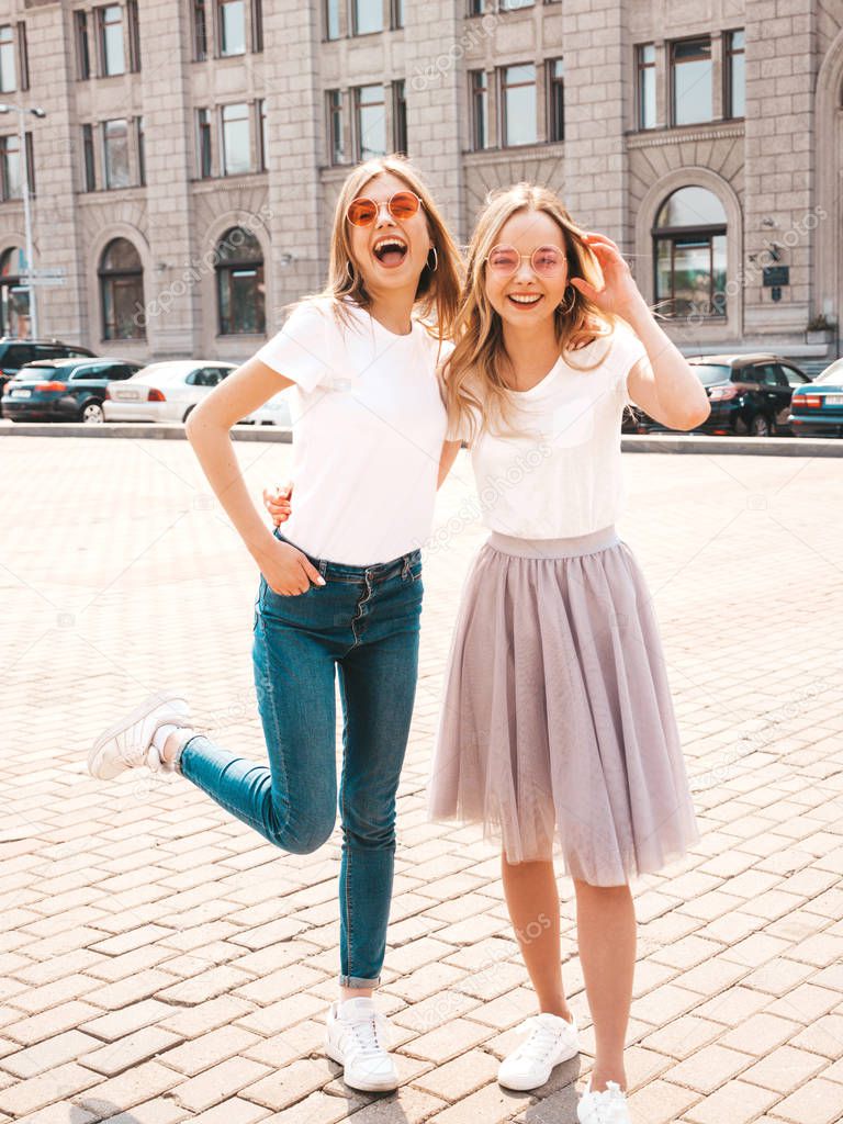 Portrait of two young beautiful blond smiling hipster girls in trendy summer white t-shirt clothes. Sexy carefree women posing on street background. Positive models having fun in sunglasses.Hugging