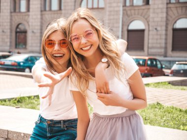 Trendy yaz beyaz t-shirt giysiler iki genç güzel sarışın gülümseyen hipster kız. Seksi kaygısız kadınlar sokak arka planda poz. Güneş gözlüğü ile eğlenen pozitif modeller. Arkadaşa ulaşmaya çalışmak