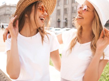 Moda yaz beyaz t-shirt giysiler iki genç güzel sarışın gülümseyen hipster kız Portresi. Seksi kaygısız kadınlar sokak arka planda poz. Güneş gözlüğü ve şapka ile eğlenen pozitif modeller