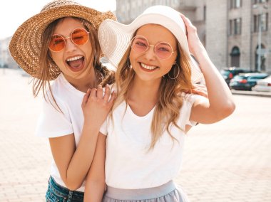 Moda yaz beyaz t-shirt giysiler iki genç güzel sarışın gülümseyen hipster kız Portresi. Seksi kaygısız kadınlar sokak arka planda poz. Güneş gözlüğü ve şapka ile eğlenen pozitif modeller