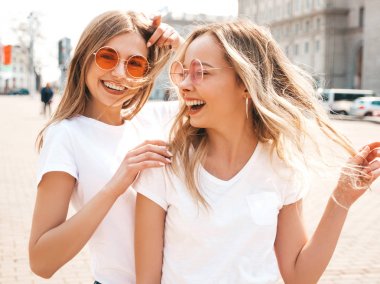 Moda yaz beyaz t-shirt giysiler iki genç güzel sarışın gülümseyen hipster kız Portresi. Seksi kaygısız kadınlar sokak arka planda poz. Güneş gözlüğü ile eğlenen pozitif modeller. Sarılma