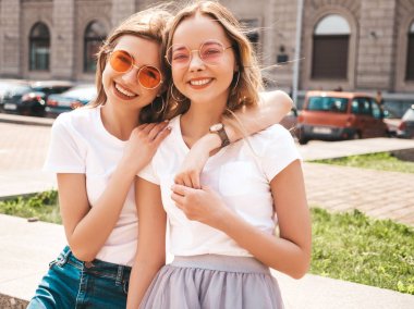 Moda yaz beyaz t-shirt giysiler iki genç güzel sarışın gülümseyen hipster kız Portresi. Seksi kaygısız kadınlar sokak arka planda poz. Güneş gözlüğü ile eğlenen pozitif modeller. Sarılma