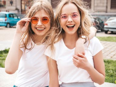 Moda yaz beyaz t-shirt giysiler iki genç güzel sarışın gülümseyen hipster kız Portresi. Seksi kaygısız kadınlar sokak arka planda poz. Güneş gözlüğü ile eğlenen pozitif modeller. Sarılma