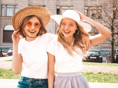 Moda yaz beyaz t-shirt giysiler iki genç güzel sarışın gülümseyen hipster kız Portresi. Seksi kaygısız kadınlar sokak arka planda poz. Güneş gözlüğü ve şapka ile eğlenen pozitif modeller