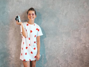 Güzel genç gülümseyen fotoğrafçı kız onu retro kamera kullanarak fotoğraf çekmek. Resim yapan kadın. Model rahat yaz beyaz t-shirt giymiş. Poz Gri duvarın yanındaki stüdyoda