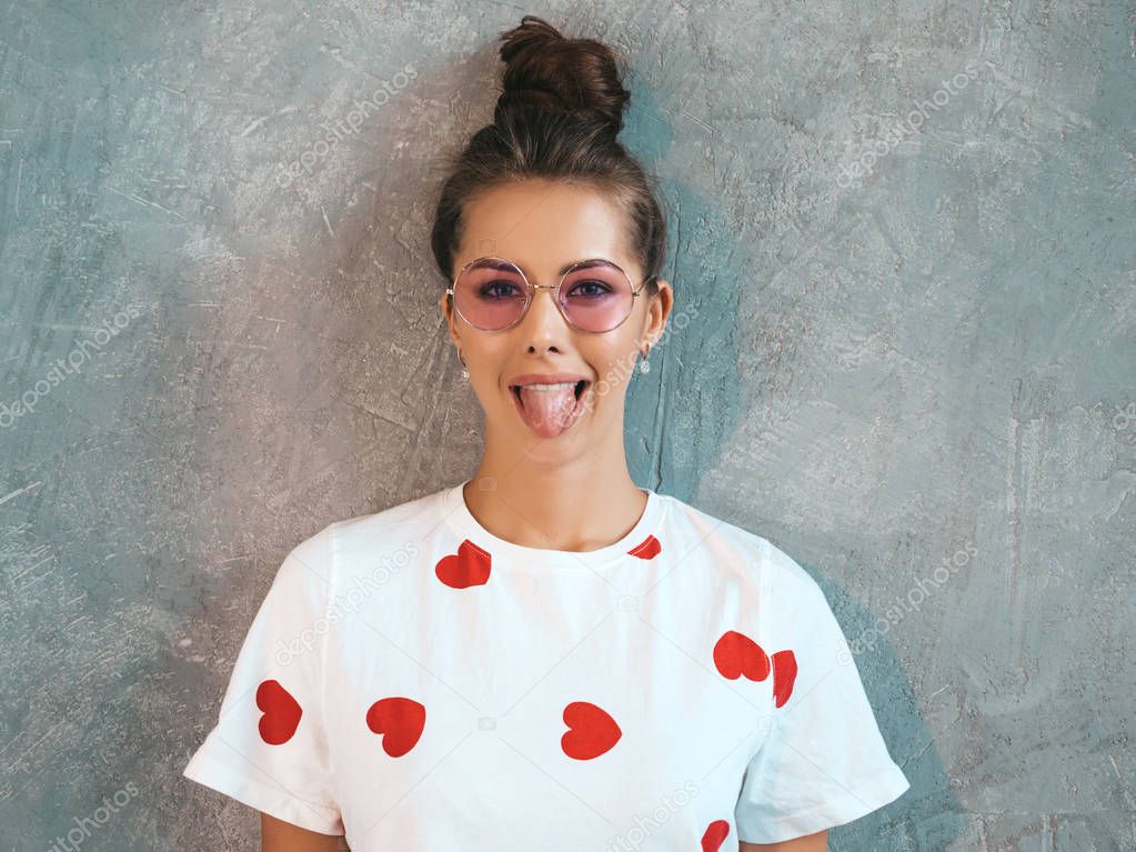 Closeup portrait of young beautiful smiling woman looking at camera. Trendy girl in casual summer white dress and sunglasses. Funny and positive female posing near gray wall in studio. Show her tongue