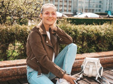 Yaz hipster ceket ve kot elbise giymiş güzel gülümseyen esmer model Portresi. Sokak arka planda bankta oturan trendy kız. Güneş gözlüğü komik ve pozitif kadın