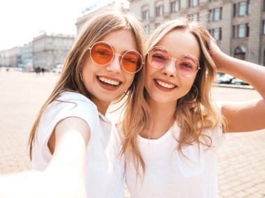 İki genç, gülümseyen, beyaz tişörtlü sarışın kadın. Akıllı telefondan selfie çeken kızlar. Arka planda poz veren mankenler. Olumlu yüz ifadeleri gösteren bir kadın.