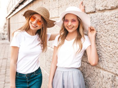 Moda yaz beyaz t-shirt giysiler iki genç güzel sarışın gülümseyen hipster kız Portresi. Seksi kaygısız kadınlar duvara yakın sokakta poz. Güneş gözlüğü ve şapka ile eğlenen pozitif modeller
