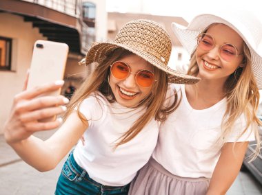 İki genç, gülümseyen, beyaz tişörtlü sarışın kadın. Akıllı telefondan selfie çeken kızlar. Arka planda poz veren mankenler. Olumlu yüz ifadeleri gösteren bir kadın.