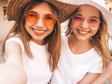 İki genç, gülümseyen, beyaz tişörtlü sarışın kadın. Akıllı telefondan selfie çeken kızlar. Arka planda poz veren mankenler. Olumlu yüz ifadeleri gösteren bir kadın.